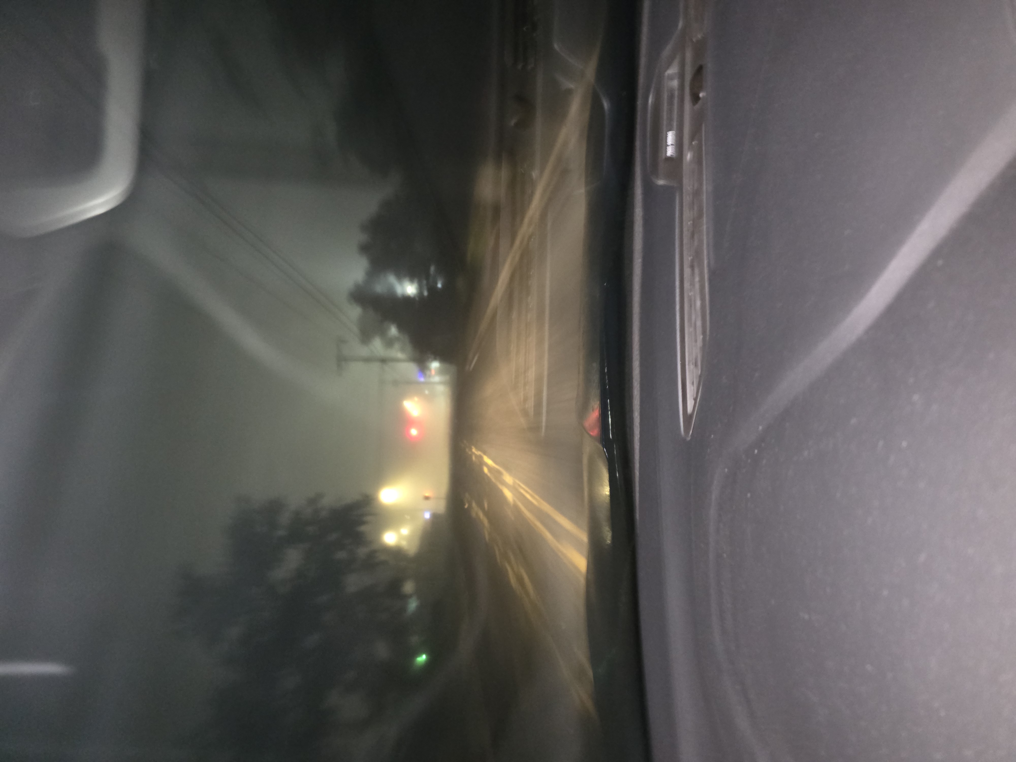 A blurry photograph taken from inside a car at night, showing a view of a road with traffic lights and faint outlines of trees in foggy or misty conditions. The dashboard of the car is visible in the foreground.
